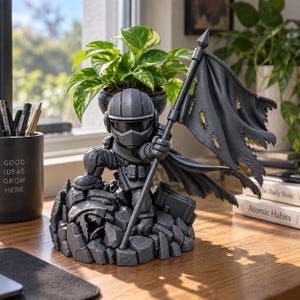 May include: A gray, stylized figurine of a soldier in tactical gear, kneeling on a rocky base while holding a large, tattered flag. The figure wears a helmet with a visor and is positioned in front of a green potted plant on a wooden desk.