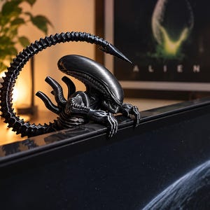 May include: A black Xenomorph figure from the Alien franchise, designed to perch on the top edge of a computer monitor. The creature features a smooth elongated head, sharp claws gripping the screen frame, and a long, segmented tail arched upward.