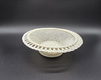 Op de afbeelding: Een ronde, gebroken witte decoratieve schaal met een ingewikkeld, open geweven roosterpatroon aan de zijkanten. De schaal heeft een brede, geschulpte rand met een gevlochten ontwerp en een stevige, geweven basis voor een gestructureerde uitstraling.