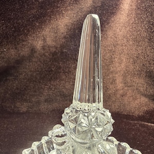 May include: A clear glass ring holder featuring a tall, faceted, tapered central post rising from a circular base. The base is decorated with a textured, diamond-cut pattern that catches the light, providing a stable foundation for the vertical display spike.