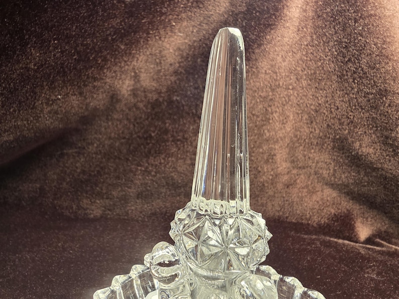 May include: A clear glass ring holder featuring a tall, faceted, tapered central post rising from a circular base. The base is decorated with a textured, diamond-cut pattern that catches the light, providing a stable foundation for the vertical display spike.