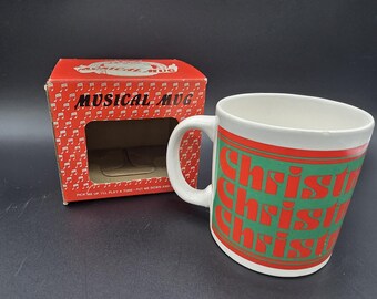 Op de afbeelding: Een witte keramische mok met een groene band waarop het woord Christmas in rode letters herhaald wordt. Daarnaast staat een rode kartonnen doos met het label Musical Mug, witte muzieknoten en de tekst Pick me up.