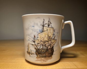 May include: A white ceramic mug featuring a detailed illustration of a large sailing ship with multiple masts and tan sails on the water. The design includes a cloudy sky background and a smaller boat in the distance, with a simple handle on the side.