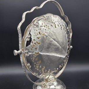 May include: A silver-plated revolving toast rack featuring an oval, pierced metal frame with intricate scrollwork and floral engravings. The central piece rotates within a decorative arched handle attached to a circular, engraved pedestal base.