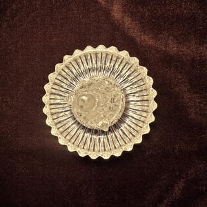 May include: A clear glass ring holder featuring a circular base with a scalloped, serrated edge and a central, faceted conical post. The glass surface displays a radial ribbed pattern on the base and a diamond-cut texture on the central post.