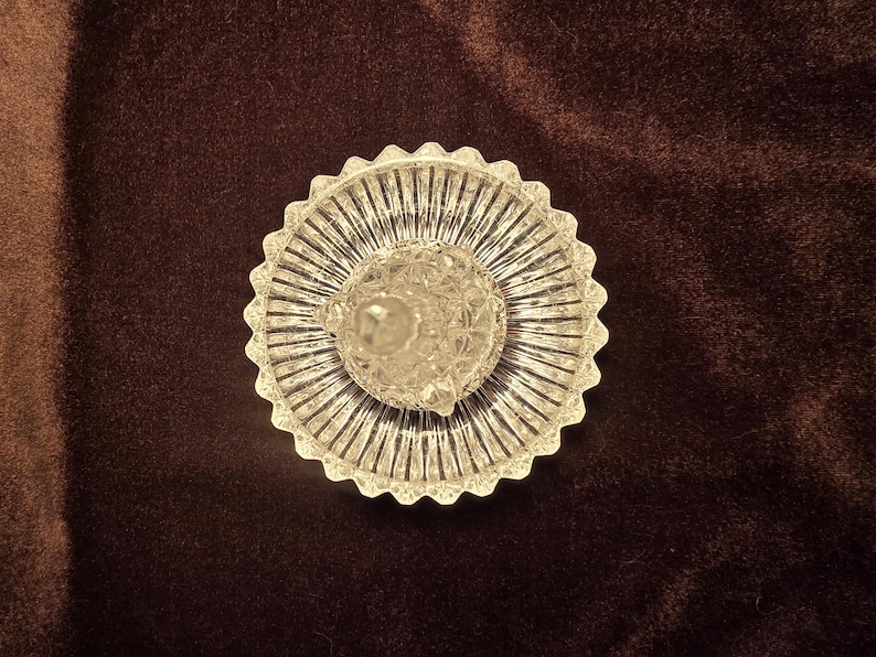 May include: A clear glass ring holder featuring a circular base with a scalloped, serrated edge and a central, faceted conical post. The glass surface displays a radial ribbed pattern on the base and a diamond-cut texture on the central post.