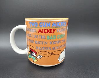 Op de afbeelding: Een keramische mok met een witte handgreep en binnenkant, met een oranje buitenkant bedrukt met de tekst TWO GUN MICKEY, OUR HERO, MICKEY LASSOS & HOG TIES THE BAD GUYS IN THIS ROOTIN' TOOTIN' 1934 WESTERN ADVENTURE en een afbeelding van Mickey Mouse.