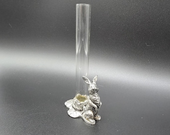 Op de afbeelding: Een vaas van een doorzichtige glazen reageerbuis die rechtop wordt gehouden door een zilverkleurige metalen voet in de vorm van een konijn dat op een blad zit. Het konijntje heeft gedetailleerde vachtstructuren en staat naast de glazen houder.