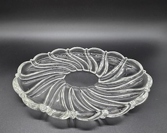 Op de afbeelding: Een serveerschaal van helder glas met een ronde opening in het midden en een geschulpte buitenrand. Het oppervlak is voorzien van een reeks verhoogde, gebogen ribbels die vanuit het midden naar buiten stralen, wat een gestructureerd waaiervormig patroon creëert.