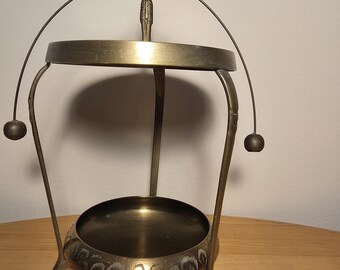 Op de afbeelding: Een messingkleurige metalen etagère met een ronde schaal aan de basis voorzien van gegraveerde bladpatronen en een bovenste ronde schaal. De constructie rust op drie gebogen poten en is bekroond met een figuur die een handvat vasthoudt.