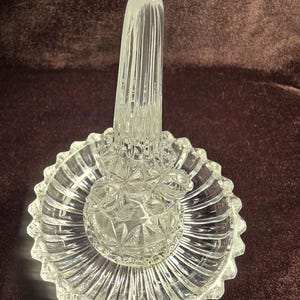 May include: A clear glass ring holder featuring a central vertical post for rings and a circular base with a scalloped edge. The glass displays a faceted, pressed pattern with geometric starburst details and vertical ribs on the base.