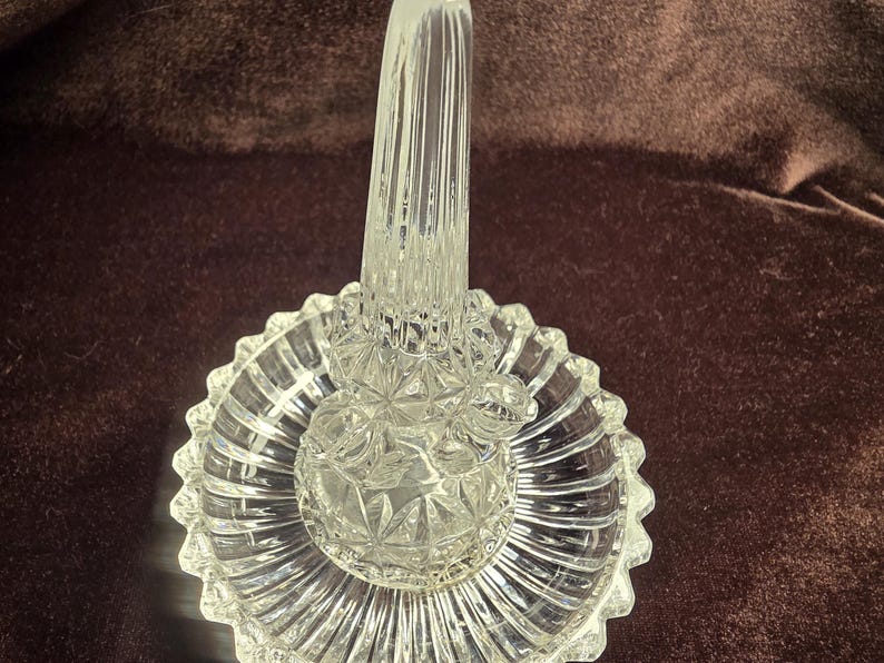 May include: A clear glass ring holder featuring a central vertical post for rings and a circular base with a scalloped edge. The glass displays a faceted, pressed pattern with geometric starburst details and vertical ribs on the base.