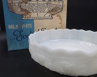 Op de afbeelding: Een witte melkglazen kom met een geschulpte rand en een reliëfpatroon van druiven en wijnranken, vergezeld door de originele kartonnen doos met het opschrift Vintage Design, W617, en Milk White Footed Bowl met een illustratie.