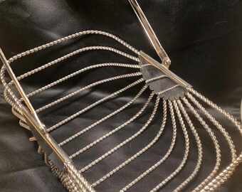 Op de afbeelding: Een rechthoekige mand van zilverkleurig metaal met een reeks gedraaide draadstrengen die de zijkanten en de bodem vormen. De mand is voorzien van een scharnierend, boogvormig handvat bevestigd aan twee waaiervormige metalen platen.
