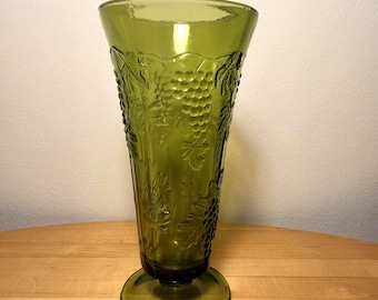 Op de afbeelding: Een hoge, olijfgroene glazen vaas met een uitlopende rand en een ronde voet. Het buitenoppervlak is versierd met een verhoogd, reliëfpatroon van druiventrossen en wijnbladeren dat rondom het gehele lichaam van de vaas is aangebracht.