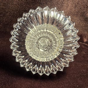 May include: A clear glass bowl featuring a starburst pattern with radiating ridges extending from a central circular base to a scalloped, serrated edge. The glass is transparent and reflective, showcasing a geometric design with sharp, defined points.