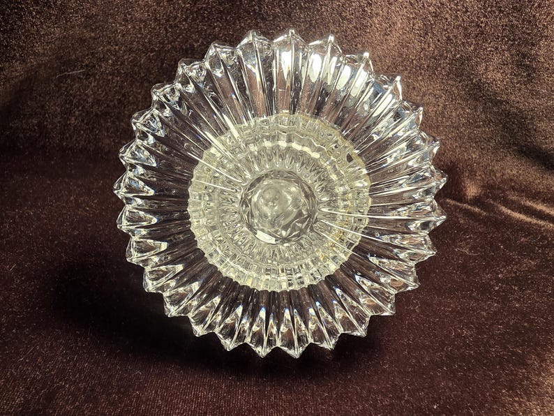May include: A clear glass bowl featuring a starburst pattern with radiating ridges extending from a central circular base to a scalloped, serrated edge. The glass is transparent and reflective, showcasing a geometric design with sharp, defined points.