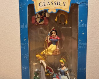 May include: A blue Disney Classics box containing miniature ornaments and package decorations. The set features figures of Snow White, Cinderella, and Aurora, along with smaller animal characters, all displayed behind a clear plastic window.