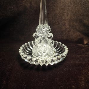 May include: A clear glass ring holder featuring a central conical post for rings, surrounded by a circular base with a scalloped edge. The base and the lower section of the post are decorated with a pressed diamond pattern, creating a faceted texture.