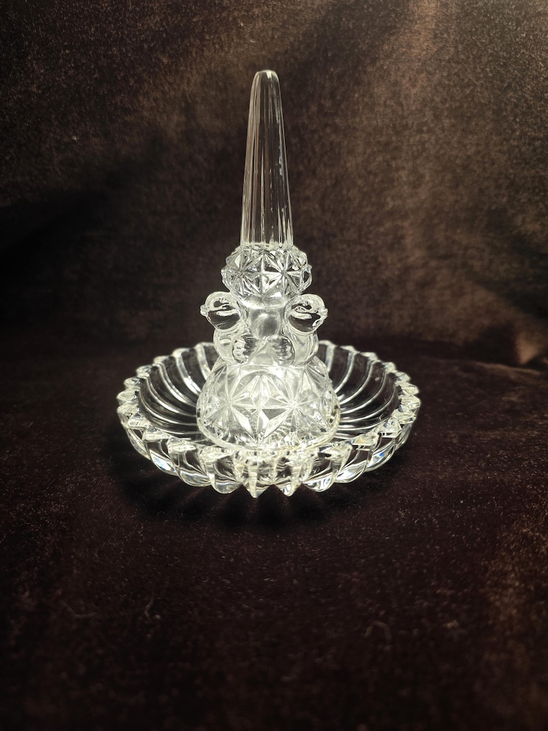 May include: A clear glass ring holder featuring a central conical post for rings, surrounded by a circular base with a scalloped edge. The base and the lower section of the post are decorated with a pressed diamond pattern, creating a faceted texture.