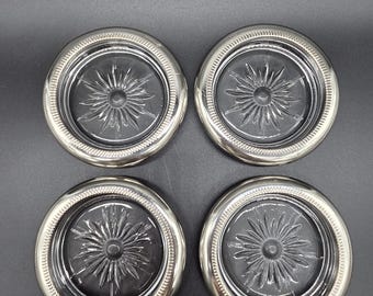 May include: A set of four circular coasters, each featuring a clear glass center with a pressed starburst pattern. The glass inserts are encased in a polished silver-tone metal rim that displays a fine, textured beaded edge detail around the border.