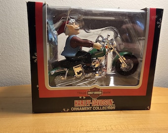 Op de afbeelding: Een Harley-Davidson ornamenten verzameldoos met een figuurtje van een personage met grijs haar en een snor op een groene motorfiets. De verpakking bevat het Harley-Davidson logo en tekst, met sneeuwvlokafbeeldingen op de zwart-rode rand.