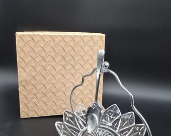 Op de afbeelding: Een heldere schaal van geperst glas met een sterpatroon op de bodem en bloembladachtige randen, voorzien van een zilverkleurig metalen handvat en een bijpassende zilverkleurige lepel, inclusief een vierkante beige doos met textuur.