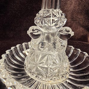 May include: A clear pressed glass centerpiece featuring a circular scalloped base with radial ribbing. The central pedestal is adorned with a geometric starburst pattern and two sculpted bird figures facing outward on opposite sides of the stem.
