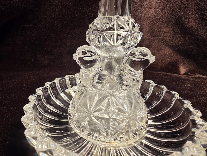 May include: A clear pressed glass centerpiece featuring a circular scalloped base with radial ribbing. The central pedestal is adorned with a geometric starburst pattern and two sculpted bird figures facing outward on opposite sides of the stem.