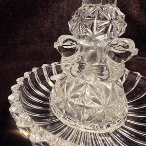 May include: A clear pressed glass centerpiece featuring a central pedestal with two sculpted bird figures and a diamond-cut pattern. The pedestal rests on a circular, scalloped-edge dish with a radial ribbed design, creating a reflective surface.