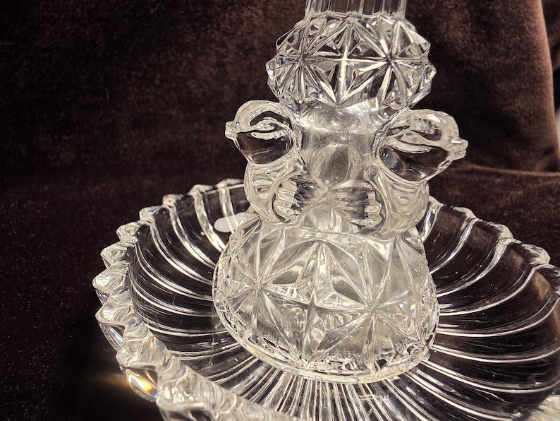 May include: A clear pressed glass centerpiece featuring a central pedestal with two sculpted bird figures and a diamond-cut pattern. The pedestal rests on a circular, scalloped-edge dish with a radial ribbed design, creating a reflective surface.