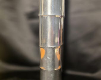 May include: A tall, cylindrical metal vase with a polished silver finish. The exterior surface features horizontal ridges that mimic the segmented appearance of a bamboo stalk, creating a smooth, reflective texture throughout the entire object.