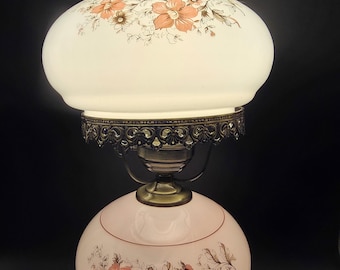 Op de afbeelding: Een tafellamp met een melkglazen kap met een gegolfde rand en een bijpassende bolvormige voet. Beide onderdelen zijn versierd met een bloemenpatroon in perzik- en bruintinten. De lamp is voorzien van een sierlijke bronzen metalen kraag.
