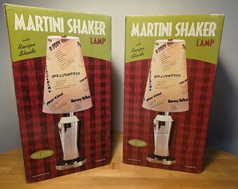 Op de afbeelding: Twee identieke productdozen voor een Martini Shaker Lamp met receptenkap. De dozen zijn groen en rood met witte tekst MARTINI SHAKER LAMP. De afbeelding toont een chromen voet en een witte kap bedrukt met verschillende cocktailrecepten.