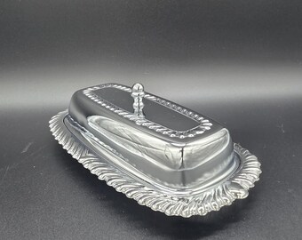 Op de afbeelding: Een rechthoekige zilverkleurige botervloot met een gepolijste, reflecterende afwerking. Het deksel heeft een kleine, ronde knop als handvat en een decoratieve kralenrand. De basis heeft een brede, geschulpte rand met gedetailleerde, stralende lijnen.