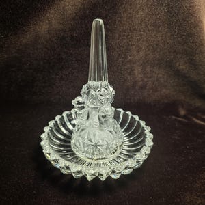 May include: A clear glass ring holder featuring a central tapered cone for rings and a circular base. The piece is crafted with a faceted, diamond-cut pattern on the cone and a scalloped, ribbed edge around the perimeter of the circular tray.