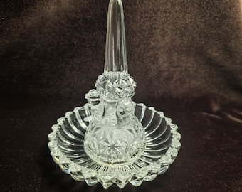 Puede incluir: Un soporte para anillos de cristal transparente con un cono central cónico para anillos y una base circular. La pieza presenta un patrón de facetas de diamante en el cono y un borde festoneado y acanalado alrededor del plato circular.