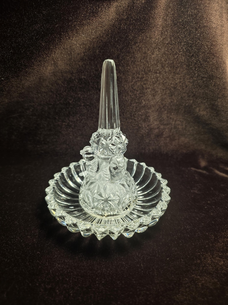 May include: A clear glass ring holder featuring a central tapered cone for rings and a circular base. The piece is crafted with a faceted, diamond-cut pattern on the cone and a scalloped, ribbed edge around the perimeter of the circular tray.