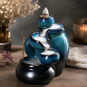 May include: A ceramic backflow incense burner featuring a tiered design with multiple rounded basins. The glaze transitions from a deep black at the base to a vibrant teal and blue at the top. White smoke cascades downwards from a lit incense cone.