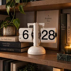 May include: A white retro-style flip clock with a central cylindrical stand and a rounded base. The clock features two white rectangular panels displaying the time in large black numerals, showing 6 PM on the left and 28 on the right.