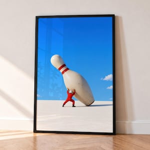 Może przedstawiać: Oprawiony wydruk artystyczny przedstawiający surrealistyczną scenę, w której mała postać w czerwonym kombinezonie i białym kasku pcha gigantyczny biały kręgiel z dwoma czerwonymi paskami na tle jasnoniebieskiego nieba i płaskiego pustynnego krajobrazu.