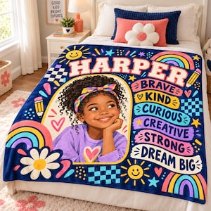 May include: A navy blue fleece blanket featuring a central illustration of a smiling child with curly hair wearing a colorful headband. The blanket includes the name HARPER, rainbows, flowers, suns, and the words BRAVE, KIND, CURIOUS, CREATIVE, STRONG, DREAM BIG.