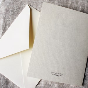 May include: A blank white card with a cream envelope. The card has a small black inscription that reads "Kayleigh DuMond".