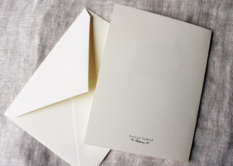 May include: A blank white card with a cream envelope. The card has a small black inscription that reads "Kayleigh DuMond".