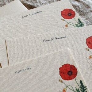 May include: Set of three blank stationery cards with a floral design. The cards are cream colored with a subtle texture. Each card features a red poppy flower with green leaves and stems. The top card has the name "Cassie T. Summers" printed in black. The middle card has the name "Cassie T. Summers" printed in black cursive. The bottom card has the words "Thank You" printed in black.