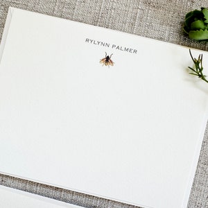 May include: White stationery card with the name "RYLYNN PALMER" printed in black at the top. A small illustration of a bee is centered below the name.