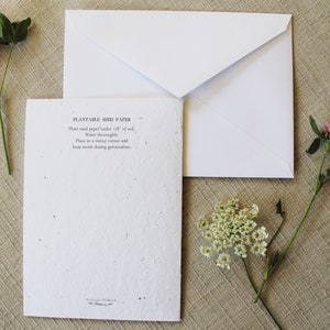 May include: A white card with the text "Plantable Seed Paper" and instructions on how to plant the paper. The card is sitting on a beige surface with a white envelope and two sprigs of flowers.