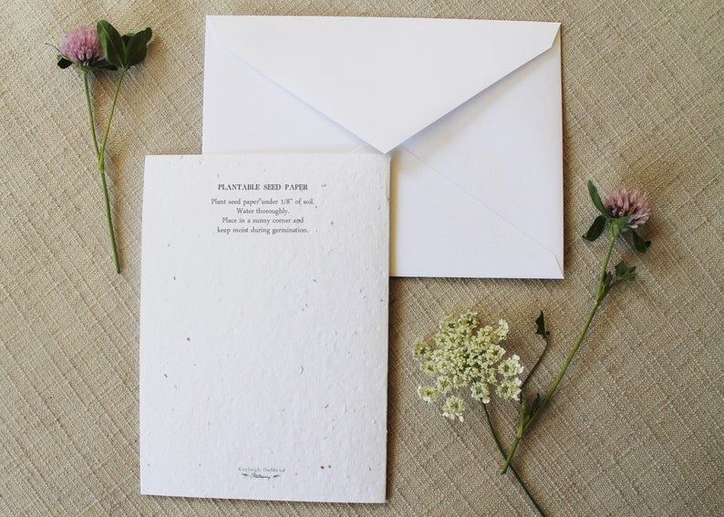 May include: A white card with the text "Plantable Seed Paper" and instructions on how to plant the paper. The card is sitting on a beige surface with a white envelope and two sprigs of flowers.