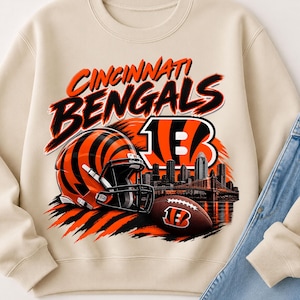 May include: A beige crewneck sweatshirt featuring a graphic print of the Cincinnati Bengals logo, a football helmet with orange and black stripes, a brown football, and a city skyline silhouette with a bridge over water in orange and black.