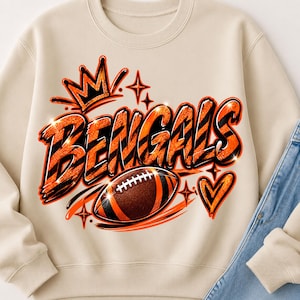 Pode incluir: Uma camisola bege de gola redonda com uma impressão gráfica que apresenta a palavra BENGALS em letras cor de laranja e pretas com riscas de tigre. O design inclui uma coroa, uma bola de futebol americano, um coração e estrelas.
